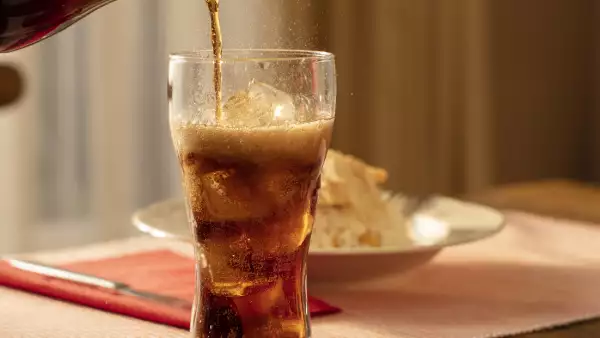 Pouring Cola from Bottle into Glass and Fizz with Ice Cubes on Table Against Blurred Livinroom  Background