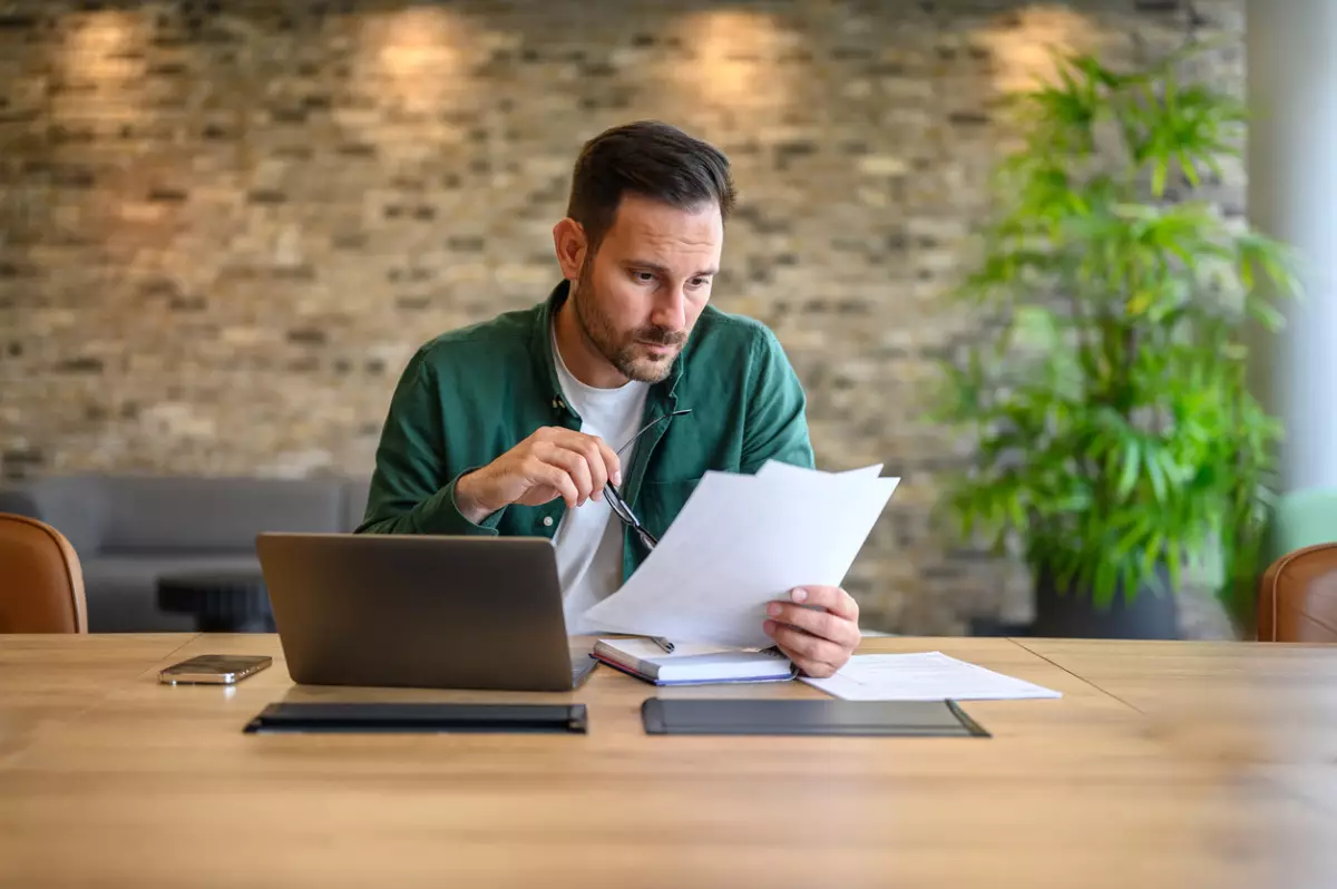 Serious male accountant holding glasses and analyzing financial report with laptop on desk in office