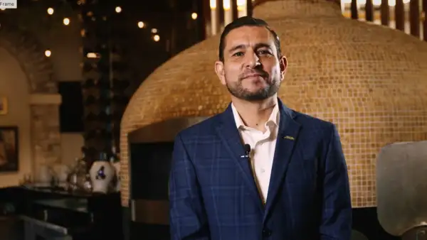 Joel Ornelas, director de Italianni’s, posa con un traje azul a cuadros y camisa blanca, frente a un horno de piedra dentro de un restaurante de la cadena.