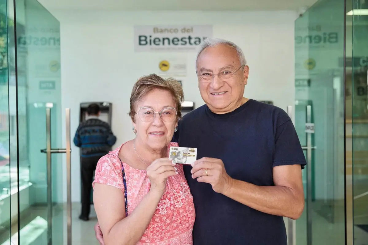 Una mujer y un hombre, ambos adultos mayores, abrazados de lado y sosteniendo una tarjeta del banco Bienestar. 