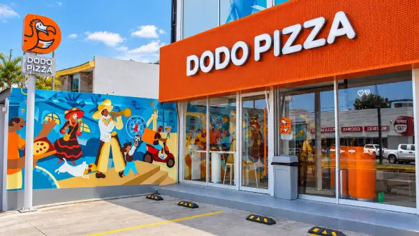 Fachada de un restaurante Dodo Pizza con letrero naranja, ventanales de cristal y un mural colorido en el exterior. Frente al establecimiento hay espacios de estacionamiento y un anuncio vertical con el logotipo de la cadena.