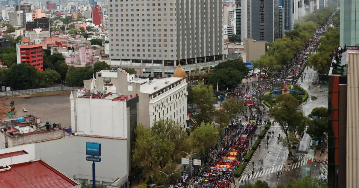 Edificios en lado izquierdo y derecho, al frente hay árboles y gente avanzando sobre una avenida, portan mantas.jpg
