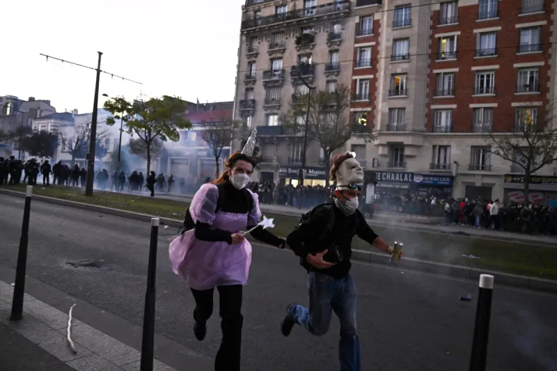 Dos manifestantes disfrazados corren mientras, al fondo, se observan nubes de gas lacrimógeno durante las protestas organizadas contra el aumento del costo de vida y la desigualdad, en París.