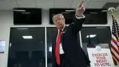 Donald Trump, con traje oscuro y corbata roja, levanta el brazo y señala hacia arriba dentro de un centro de votación en Florida. A su lado hay un cartel que invita a ejercer el derecho al voto y una bandera de Estados Unidos.