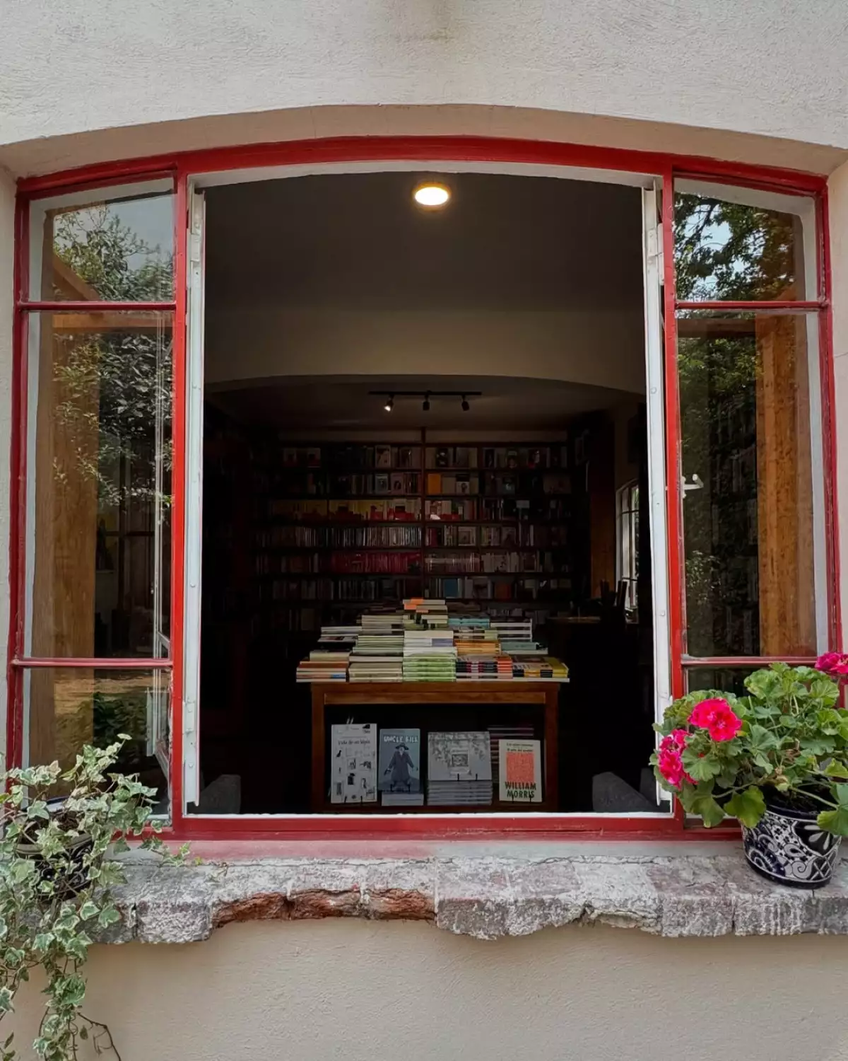 Foto de una ventana con marco color rojo y flores en la cornisa, hacia adentro una sala llena de libros.
