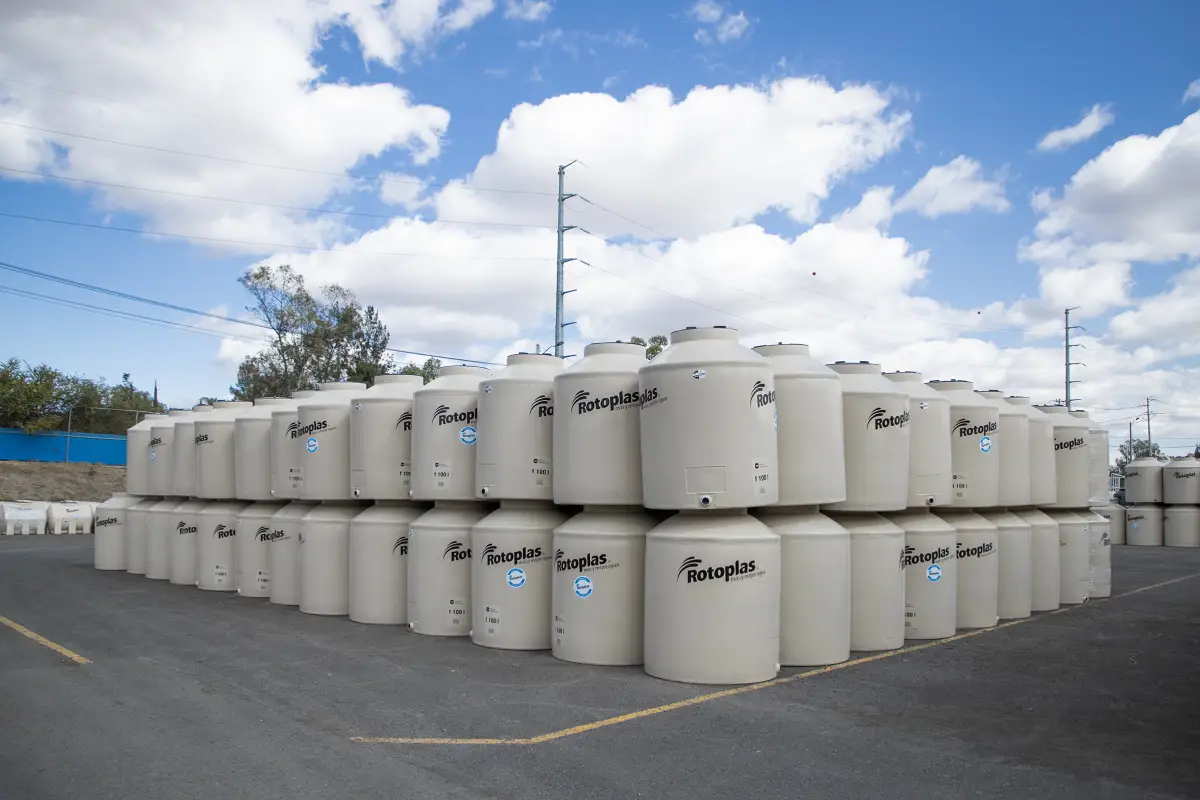 Un espacio exterior de almacenamiento donde se observan decenas de grandes tanques cilíndricos blancos de almacenamiento de agua, colocados en varias hileras y niveles. Los depósitos muestran el logotipo de Rotoplas y una capacidad visible de 1,100 litros. Los tanques ocupan la mayor parte de la imagen y están sobre un amplio pavimento oscuro con líneas amarillas, mientras que al fondo se distinguen árboles, una barda azul, postes y cables eléctricos. El cielo azul intenso, cubierto parcialmente por grandes nubes blancas, contrasta con los depósitos y el entorno industrial.
