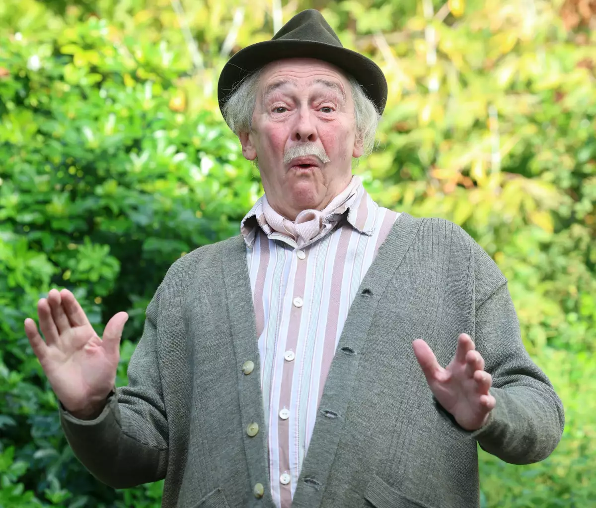 Foto del actor Paul Whitehouse con gesto de sorpresa, viste sombrero, suéter gris de botones y camisa a rayas.