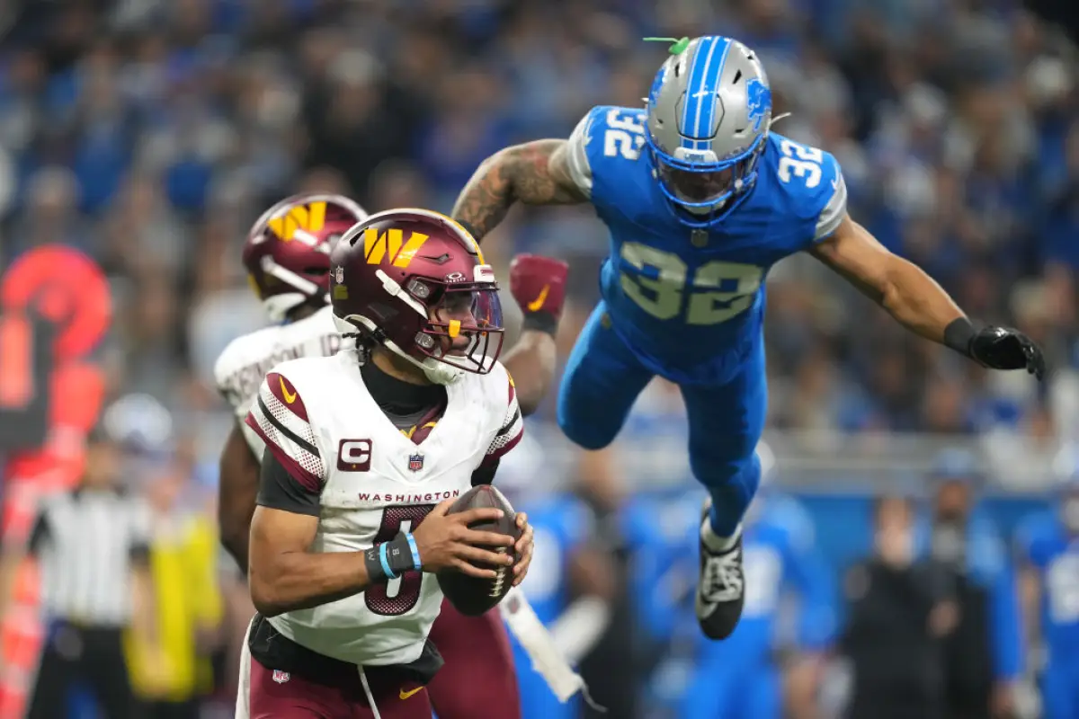 Foto de Jayden Daniels, de los Commanders, corre con el balón en mano, mientras que detrás "volando" se ve a Brian Branch de los Detroit Lions intentando detenerlo.