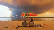 Los bañistas se sientan en la playa de Moutchic en Lacanau, al suroeste de Francia, el 24 de julio de 2026, mientras las nubes de humo se elevan al cielo a partir de un incendio forestal.