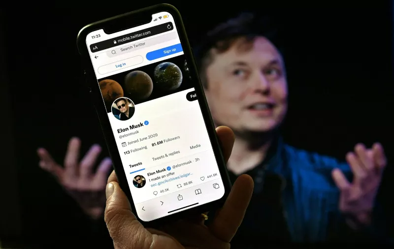 Elon Musk pausa la compra de Twitter. (Elon Musk al fondo y en primer plano un smartphone con la app de Twitter abierta). 
