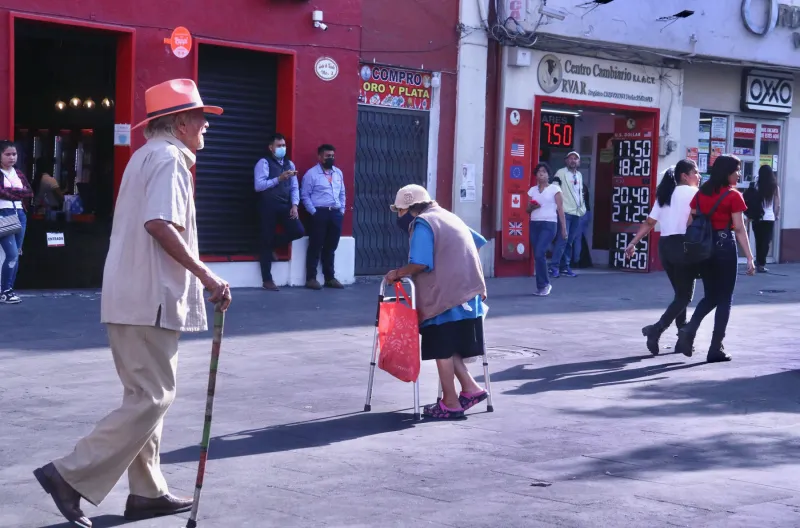 Dos personas de la tercera edad caminan apoyados en un bastón y una andadera en una calle del centro de Cuernavaca.