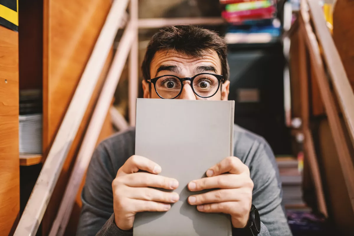 Handsome caucasian man with eyeglasses covering his face with book and looking at camera. Bookstore interior.