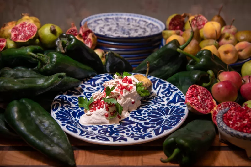 Foto de un plato de talavera poblana con un chile en nogada tradicional, rodeado de chiles poblanos crudos, frutas de temporada y granadas rojas abiertas
