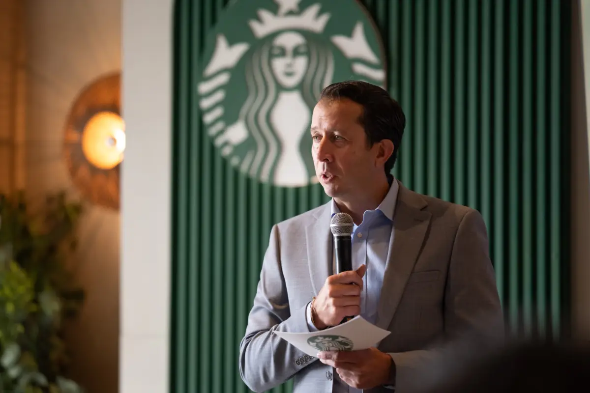 Jaime Vásquez, director de Starbucks, en traje gris con camisa azul. Sostiene un micrófono en la mano izquierda y una tarjeta en la mano derecha. Al  fondo está una pared color verde en la que sobre sale el logitipo de Starbucks, que es una sirena con dos colas en color verde.