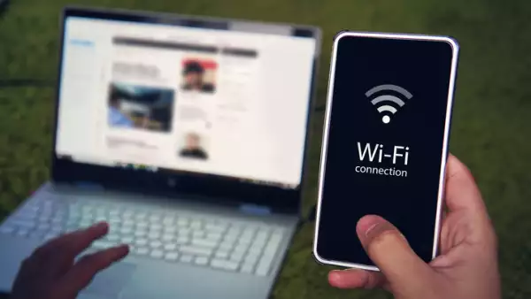 Smartphone with wifi connection generic icon on the screen hold by man on the background of laptop display and keyboard. Man uses Wi-Fi hot spot to get to domestic internet access. Wireless gadgets