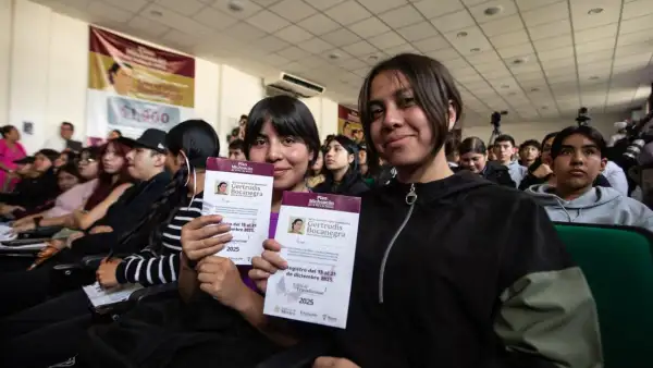 Dos estudiantes de preparatoria sentadas y rodeadas de más estudiantes muestran la tarjeta en la que reciben el pago de la beca Benito Juárez.