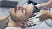 Foto de un hombre joven con los ojos cerrados mientras le lavan el cabello en una peluquería con espuma abundante.
