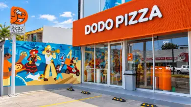 Fachada de un restaurante Dodo Pizza con letrero naranja, ventanales de cristal y un mural colorido en el exterior. Frente al establecimiento hay espacios de estacionamiento y un anuncio vertical con el logotipo de la cadena.
