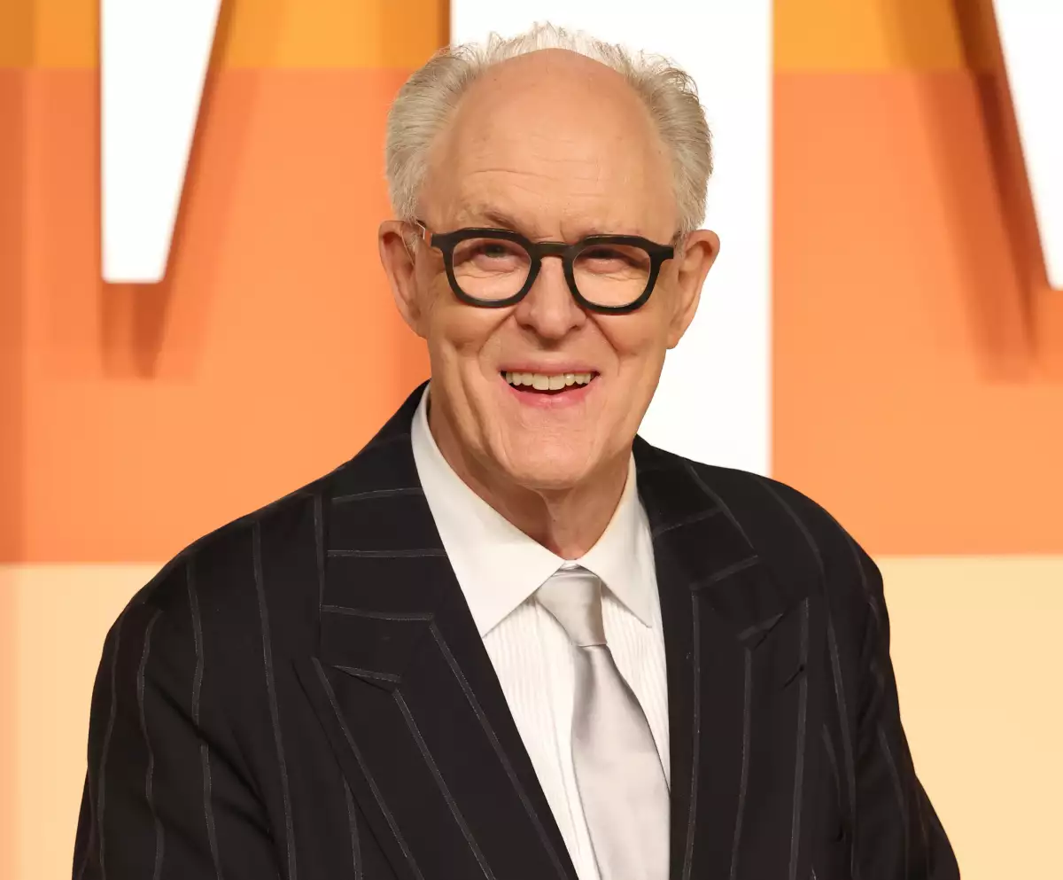 Foto del actor John Lithgow vestido de saco oscuro, camisa blanca, corbata plateada y lentes de pasta negros.