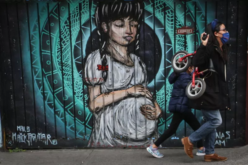 Una madre carga la bicicleta de su hija, mientras ella camina a su lado, frente a un mural de una mujer embarazada.