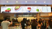 Mostrador de Chipotle con una fila de clientes que esperan para ordenar sus alimentos.
Al fondo, varios empleados preparan los pedidos detrás de una barra de servicio.
Sobre el mostrador se observa un menú con opciones de bowls, burritos, tacos y ensaladas.
A un costado hay estantes con bolsas de chips y otros complementos.