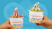 Dos vasos de helado tipo yogurt congelado de Nutrisa, sostenidos por manos desde los extremos izquierdo y derecho de la composición. Ambos vasos son blancos y llevan el logotipo de Nutrisa junto con un diseño multicolor de figuras y frutas.
