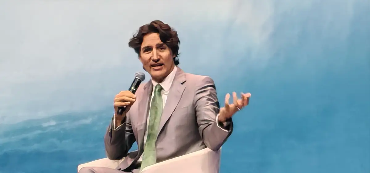 Justin Trudeau, exprimer ministro de Canadá, habla durante un evento sentado en un sillón blanco, con traje gris claro, camisa blanca y corbata verde. Sostiene un micrófono y gesticula con la mano izquierda frente a un fondo azul.
