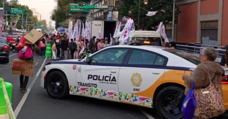 Una patrulla color blanco con vivos amarillos con letras que dice policía y tránsito en una calle con personas y pancartas.jpg