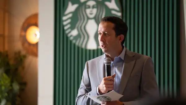 Jaime Vásquez, director de Starbucks, en traje gris con camisa azul. Sostiene un micrófono en la mano izquierda y una tarjeta en la mano derecha. Al  fondo está una pared color verde en la que sobre sale el logitipo de Starbucks, que es una sirena con dos colas en color verde.