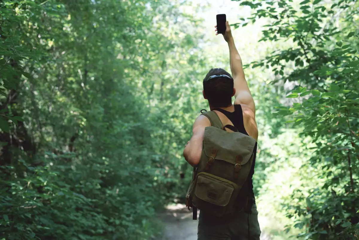 Foto de un hombre caminando por el bosque y levantando su celular con el brazo para intentar obtener conexión.