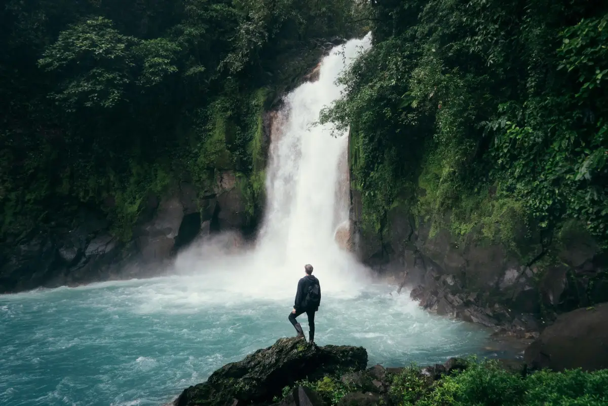 Foto de un hombre parado sobre unas rocas frente a una cascada en el bosque.