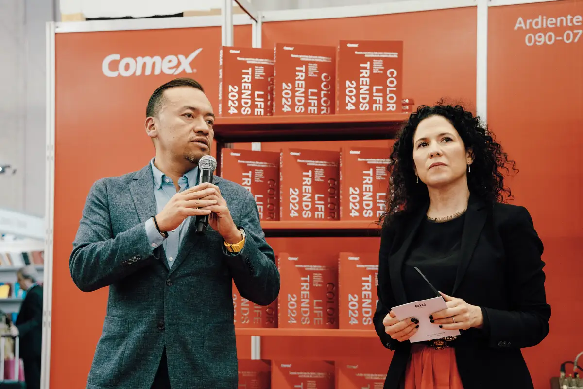 Hombre  vestido de gris y mujer vestida de negro, en el fondo se ven unos libros de la marca Comex en color naranja.
