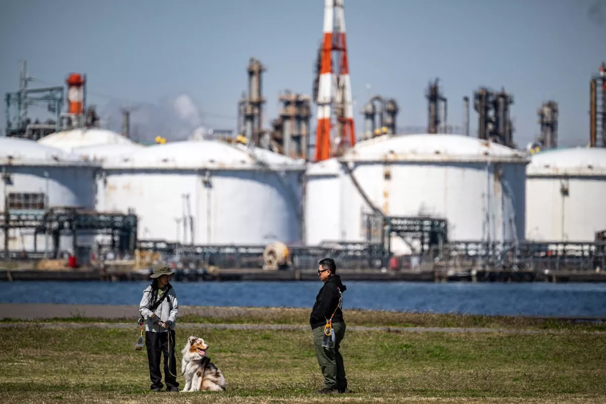 La gente juega con un perro en el Parque Higashi-Ogishima Higashi, ya que se ven tanques de almacenamiento de energía cerca de una refinería de petróleo en la Zona Industrial de Keihin, un importante cinturón industrial de la Bahía de Tokio, en la ciudad de Kawasaki el 15 de marzo de 2026.