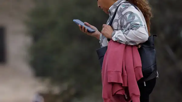 Una mujer en la calle caminando mientras mira su celular con la mano.