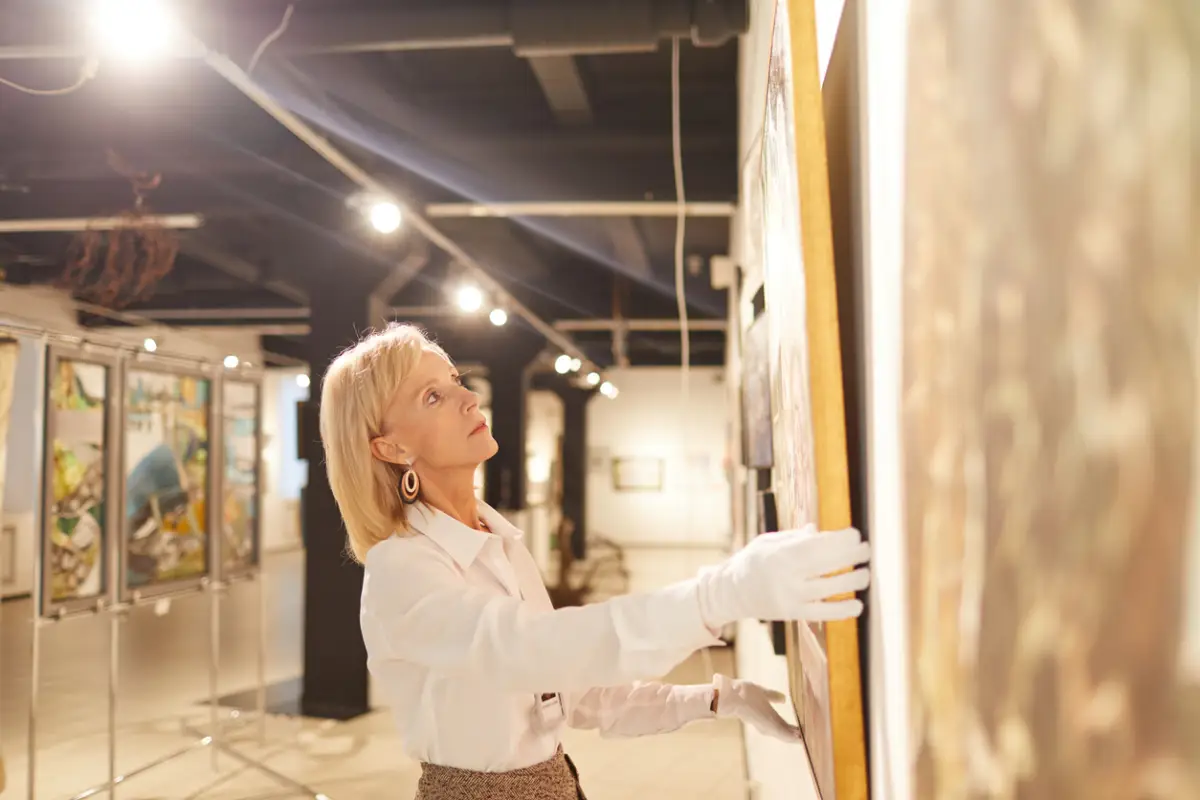 Una galerista coloca una pintura en la pared al interior de una sala de arte.