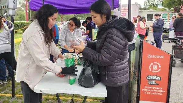 Dos mujeres frente a frente en una carpa al aire libre, una revisa con guantes un recipiente. Otra con chamarra negra lo entrega.jpg