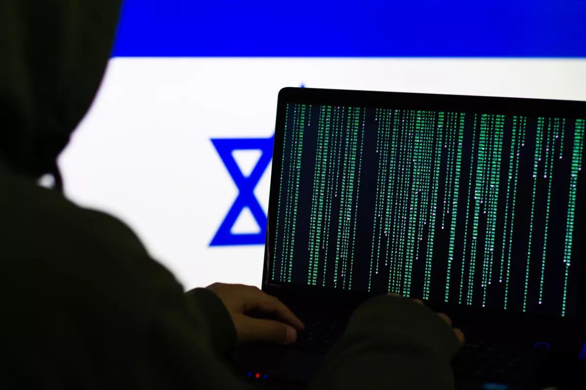 Hacker escribiendo código en una laptop con la bandera de Israel de fondo. 