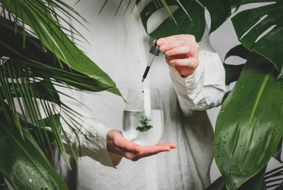 woman in a white shirt adds drops of chlorophyll to glass of water standing next to palm trees