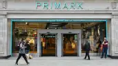 Entrada de una tienda Primark, en la que se muestran aparadores con ropa y zapatos de colores neutros como gris y nude. La fachada es color azul, con el nombre de la cadena en el mismo color en la parte de arriba de la entrada.

