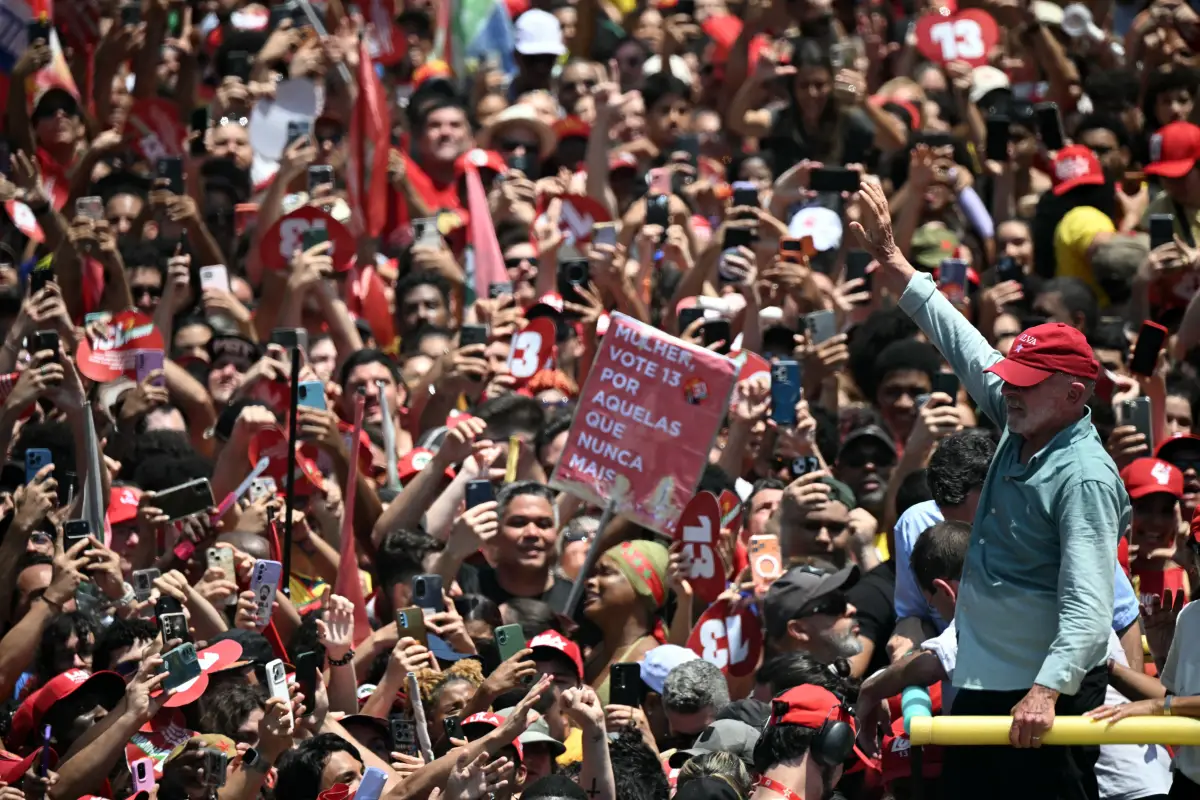 El presidente Luiz Inácio Lula da Silva saluda a una multitud de simpatizantes durante un acto de campaña en Brasil. Los asistentes, vestidos principalmente de rojo, levantan sus teléfonos para fotografiarlo y portan banderas y carteles con el número 13, asociado a su candidatura. Lula viste una camisa azul claro y una gorra roja mientras avanza entre la concentración.