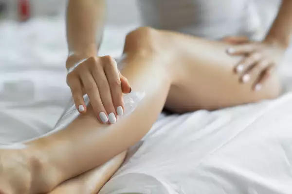 Woman applying body lotion on her legs at her bedroom.