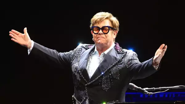 Foto de Elthon John en el escenario, con una chaqueta brillante, camisa blanca y lentes oscuros.