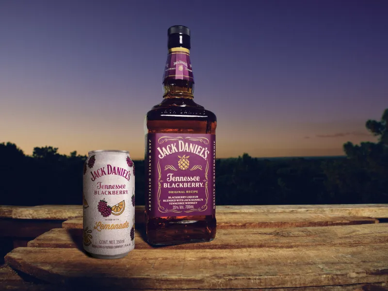 Botella de Jack Daniel’s Tennessee Blackberry y lata de Jack Daniel’s Blackberry Lemonade sobre una superficie de madera, con un paisaje arbolado y el cielo al atardecer como fondo.
