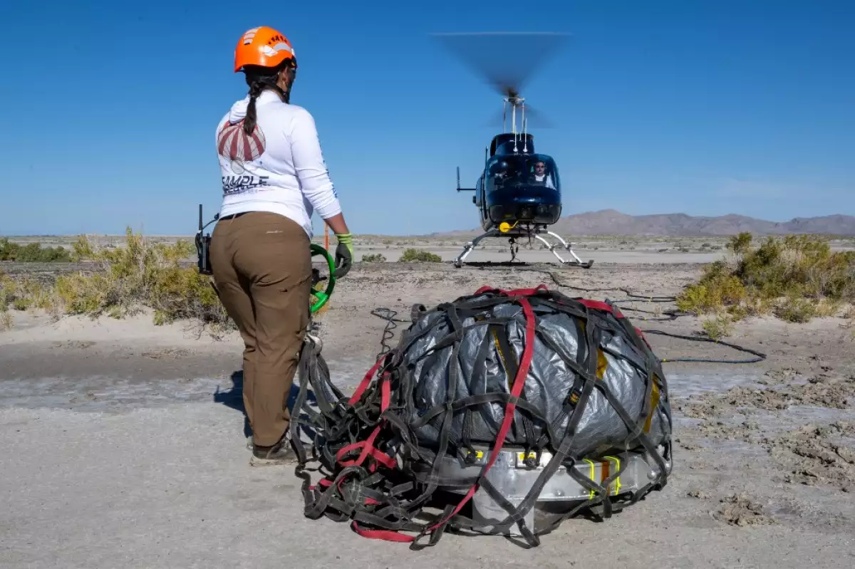 En la escena, la comandante de recuperación Jasmine Nakayama coloca la cápsula de retorno de muestras de la misión OSIRIS-REx de la NASA a un helicóptero para su transporte a la sala limpia el 24 de septiembre de 2023, poco después de que la cápsula aterrizara en el campo de pruebas y entrenamiento de Utah del Departamento de Defensa en Dugway, Utah.