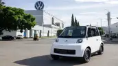 Vehículo eléctrico Olinia de color blanco, modelo mexicano, dentro del complejo industrial de la planta de Volkswagen de México en Puebla, con el edificio de producción, el logotipo de Volkswagen en la parte superior, árboles verdes y patio industrial soleado.