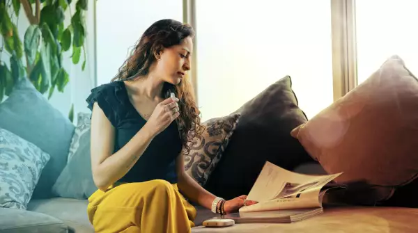 Una mujer está usando un dispositivo IQOS, mientras lee un libro, sentada en el sillón de su casa. 