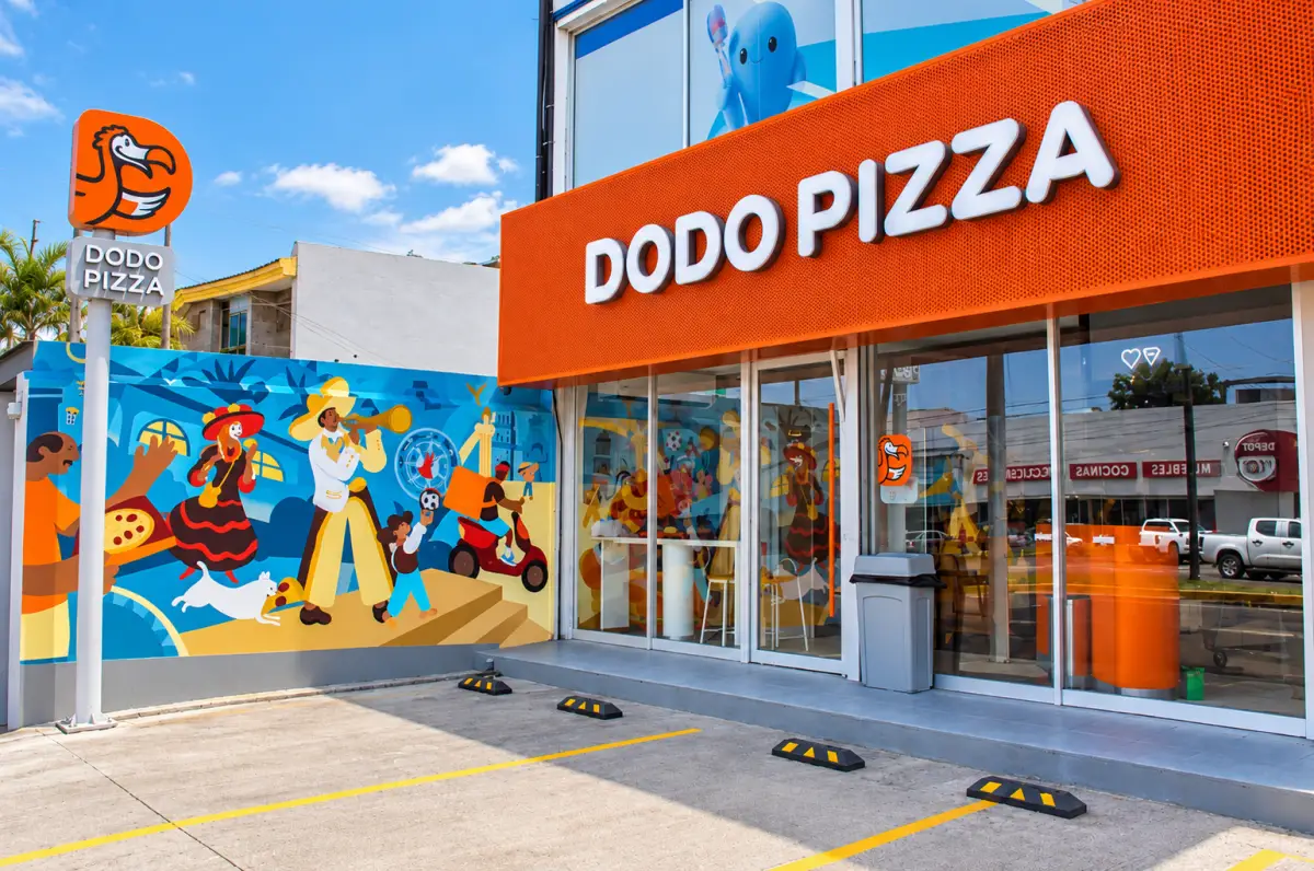 Fachada de un restaurante Dodo Pizza con letrero naranja, ventanales de cristal y un mural colorido en el exterior. Frente al establecimiento hay espacios de estacionamiento y un anuncio vertical con el logotipo de la cadena.
