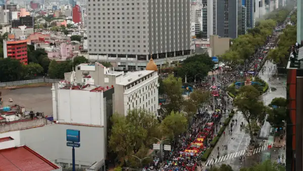 Edificios en lado izquierdo y derecho, al frente hay árboles y gente avanzando sobre una avenida, portan mantas.jpg