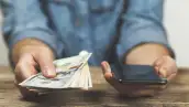 Una persona sostiene en una mano unos dólares y con la otra revisa su teléfono móvil.