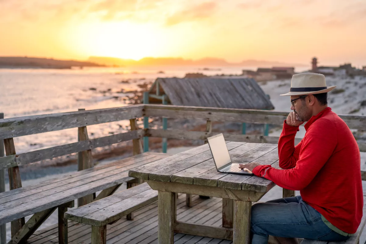 Nómada digital sentado al aire libre en la playa con un portátil solo haciendo teletrabajo al atardecer